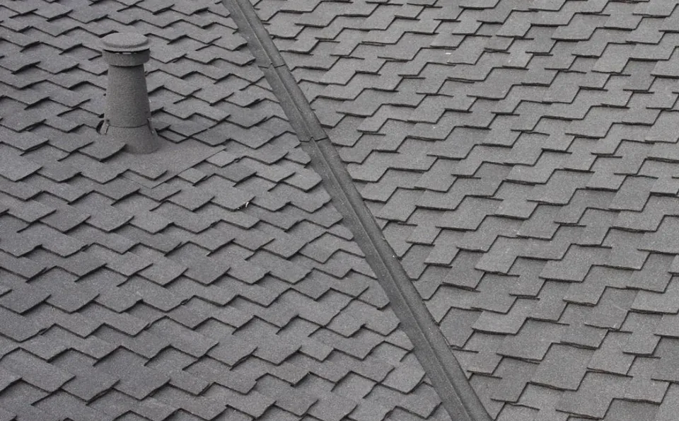Close-up of dark grey scalloped asphalt shingles with a plain grey pipe vent and a ridge cap seam running diagonally across the roof